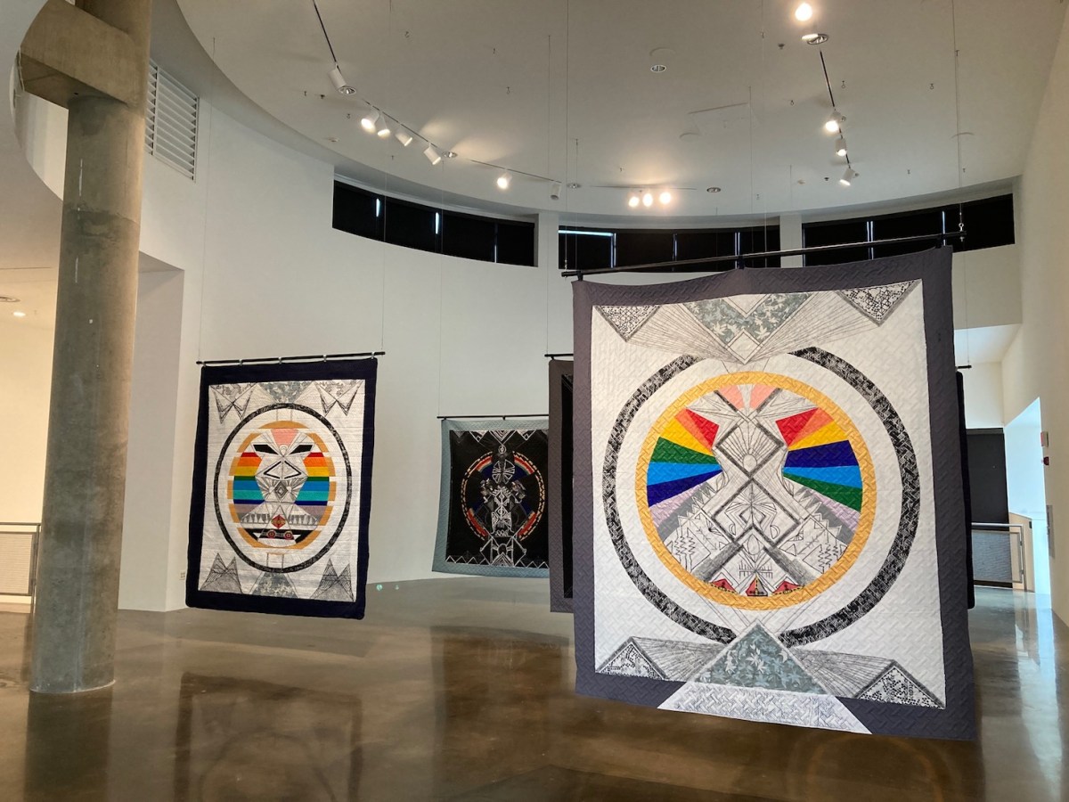 In a modern gallery space with shiny concrete floors and high vaulted ceilings, about four large tapestries, all resembling quilts, float in space. They are hung from the ceiling by thin, invisible wire. Each tapestry features a circular geometric central design that is filled with abstract patterns. Along the top and bottom of the tapestries, pyramids point up and down, somewhat resembling mountains.