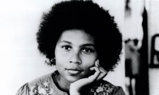 A black-and-white portrait photograph of a dark-skinned woman with a medium-sized afro. She rests her chin on her left hand and stares at the camera with a kind, soft expression. She wears an embroidered floral blouse.