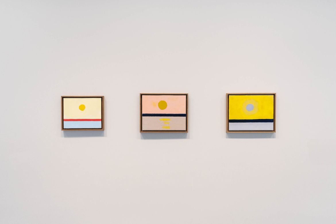 Three small paintings hang in a row on a white wall. They each feature a circle resembling a sun, a middle line resembling a horizon, and a lower section resembling the ocean. The colors differ slightly, with one sky being bright yellow, one pink, and the other a muted orange. The ocean water also differs slightly, as does the horizon line, which is red in the first painting and navy blue in the others.