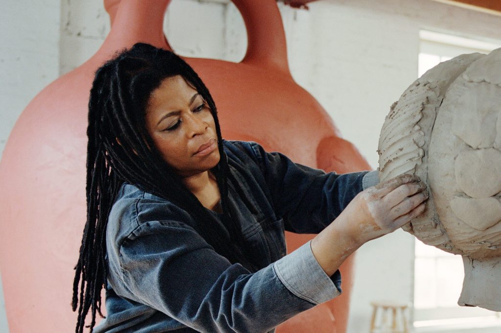 A dark-skinned woman with long black dreadlocks is pictured from the waist up working on a large circular sculpture, seemingly made from clay. She stares intent on her work and her hands are covered in the dried, chalky substance. Behind her stands a large sculpture in an earthy orange color.