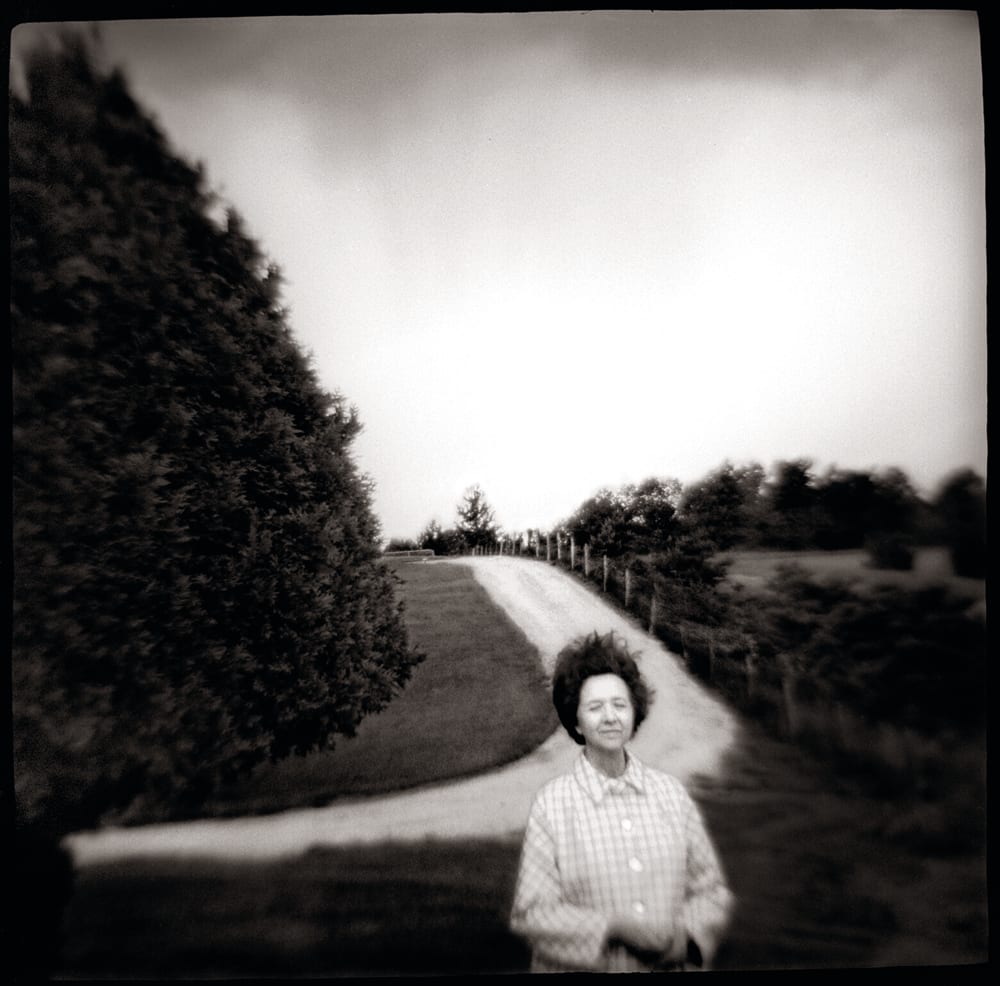 In this black-and-white photo, an older light-skinned woman is pictured in the lower frame, her short brown hair blown up by the wind. She seems to smile slightly, her eyes are closed, and her hands are clasped pleasantly in front of her stomach. She wears a 1950s-style button-up plaid shirt. Behind her is a rolling, rural landscape, including a long dirt road, a large tree in a triangle shape, and fence posts and other brush.