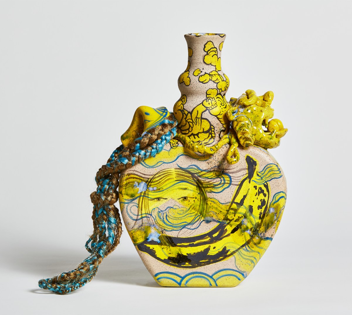 A colorful ceramic vase features abstract painted illustrations of a Asian woman's face, a ripe banana, and other lines and forms in yellow and blue. The narrow neck of the vase has braided synthetic hair, also in blue and yellow, draped and knotted around it.
