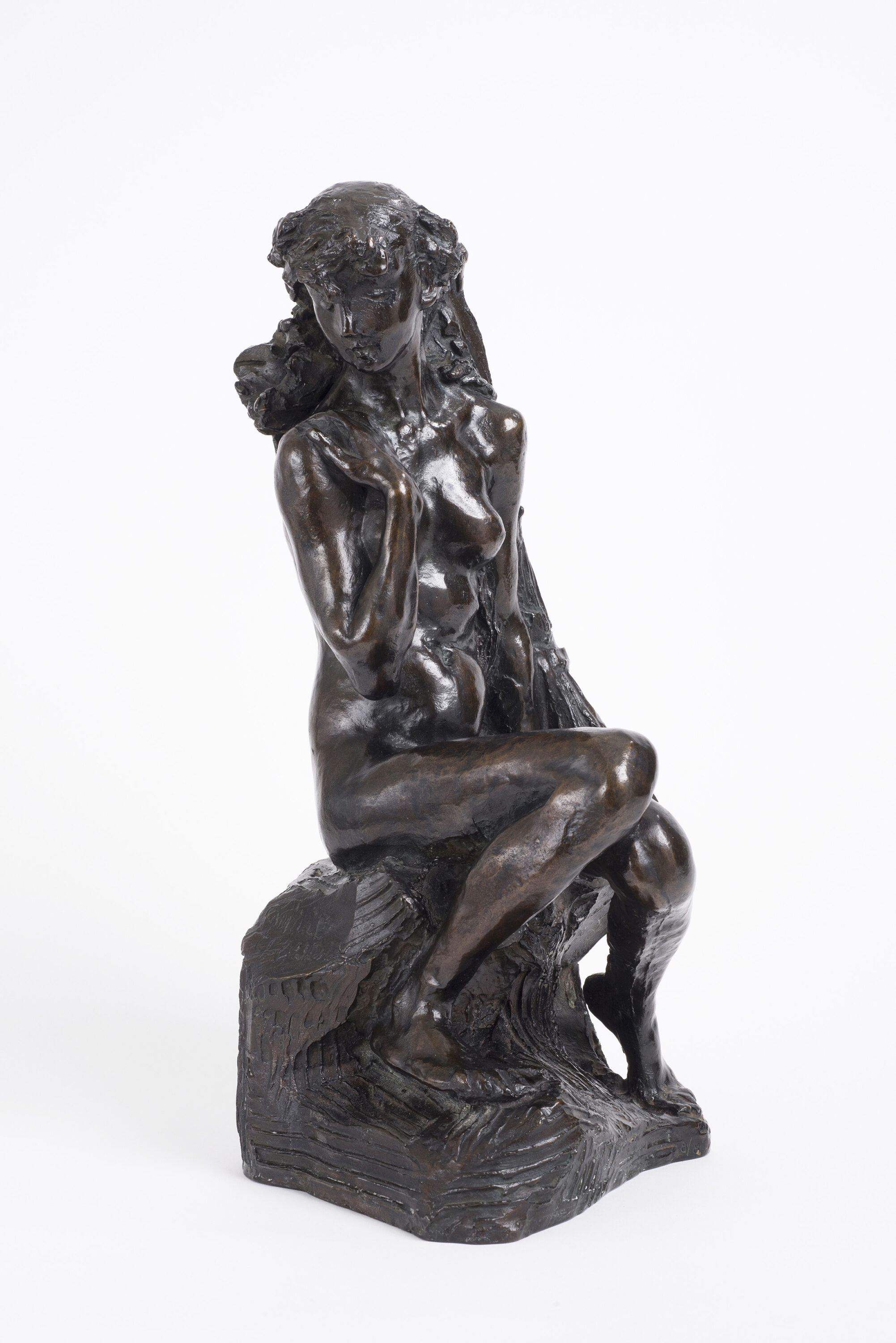 Bronze tabletop sculpture depicting a nude young woman seated on rough-hewn base, leaning against a sheaf of wheat. The figure's knees are drawn together, her left arm hanging at her side and her right arm bent upwards, clasping her shoulder.