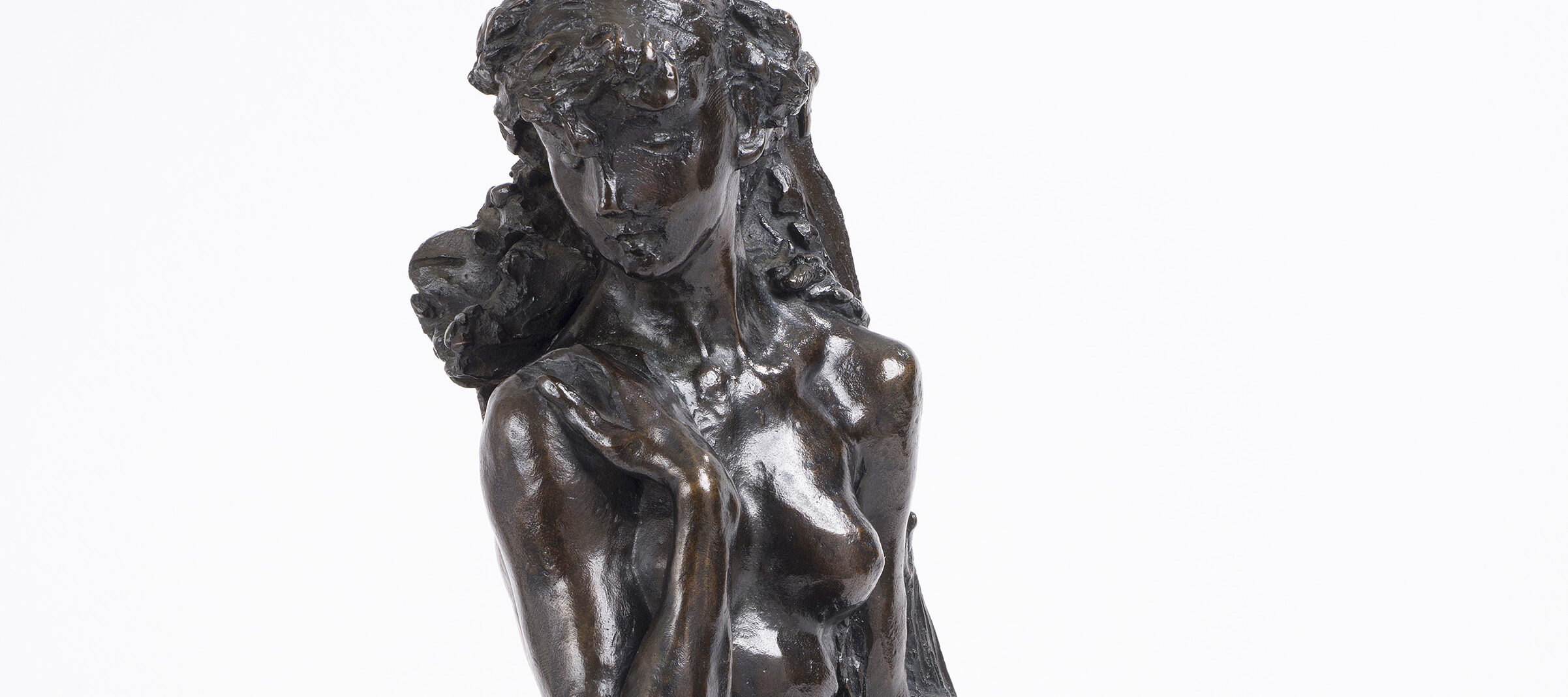 Bronze tabletop sculpture depicting a nude young woman seated on rough-hewn base, leaning against a sheaf of wheat. The figure's knees are drawn together, her left arm hanging at her side and her right arm bent upwards, clasping her shoulder.