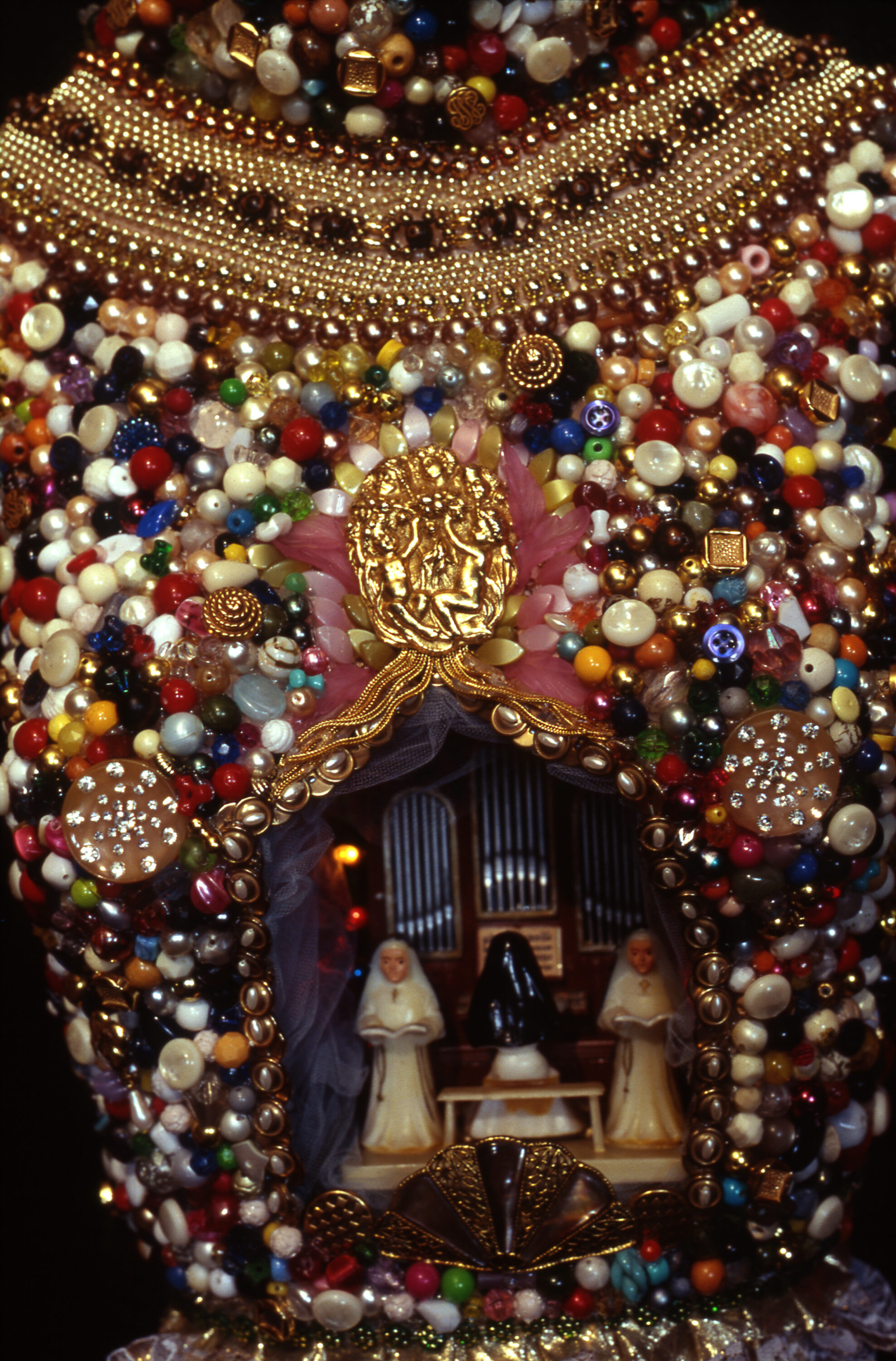 A hole in colorful beading with figures inside. The figures are a person in a veil and long dress sitting at an organ with two light-skinned people in the same attire holding open Bibles on either side of the organ.