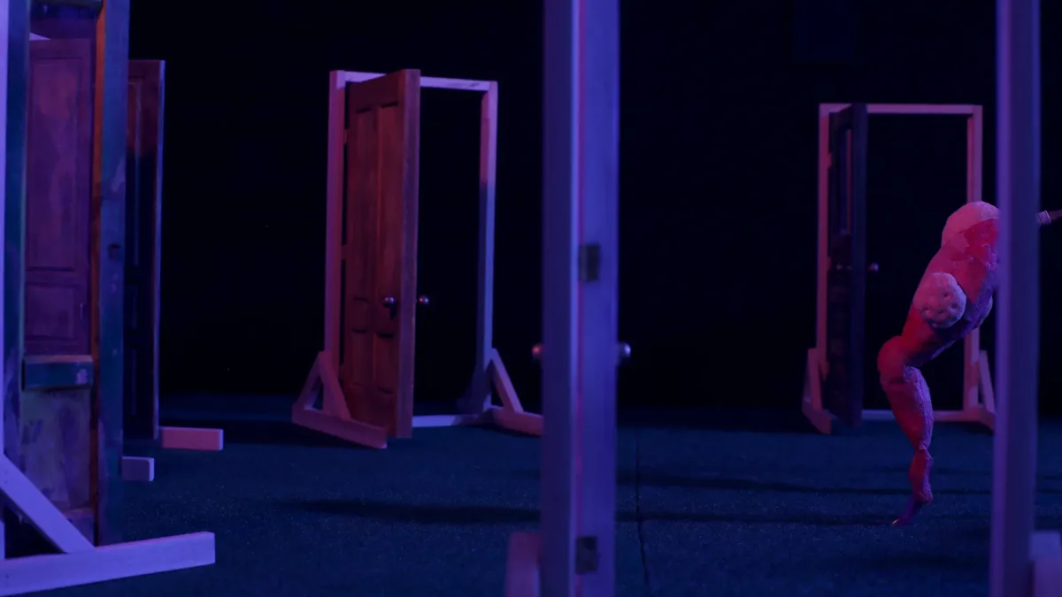 In this screen still from a puppet stop motion animation, four minature doors set in wooden frames are arranged in a dark room, lit in shades of deep purple and red. A strange, abstract wooden figure stands on one leg to the right, almost out of frame.