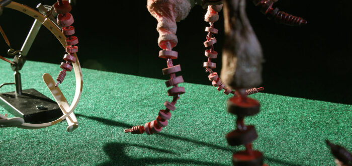 In this screen still from a puppet stop motion animation, miniature sculptures made from wood, metal, and other materials are arranged on top of, and hang above, green turf, set against a pitch black background. The sculpture's shadows appear on the turf. The sculptures are made up of various circular designs.