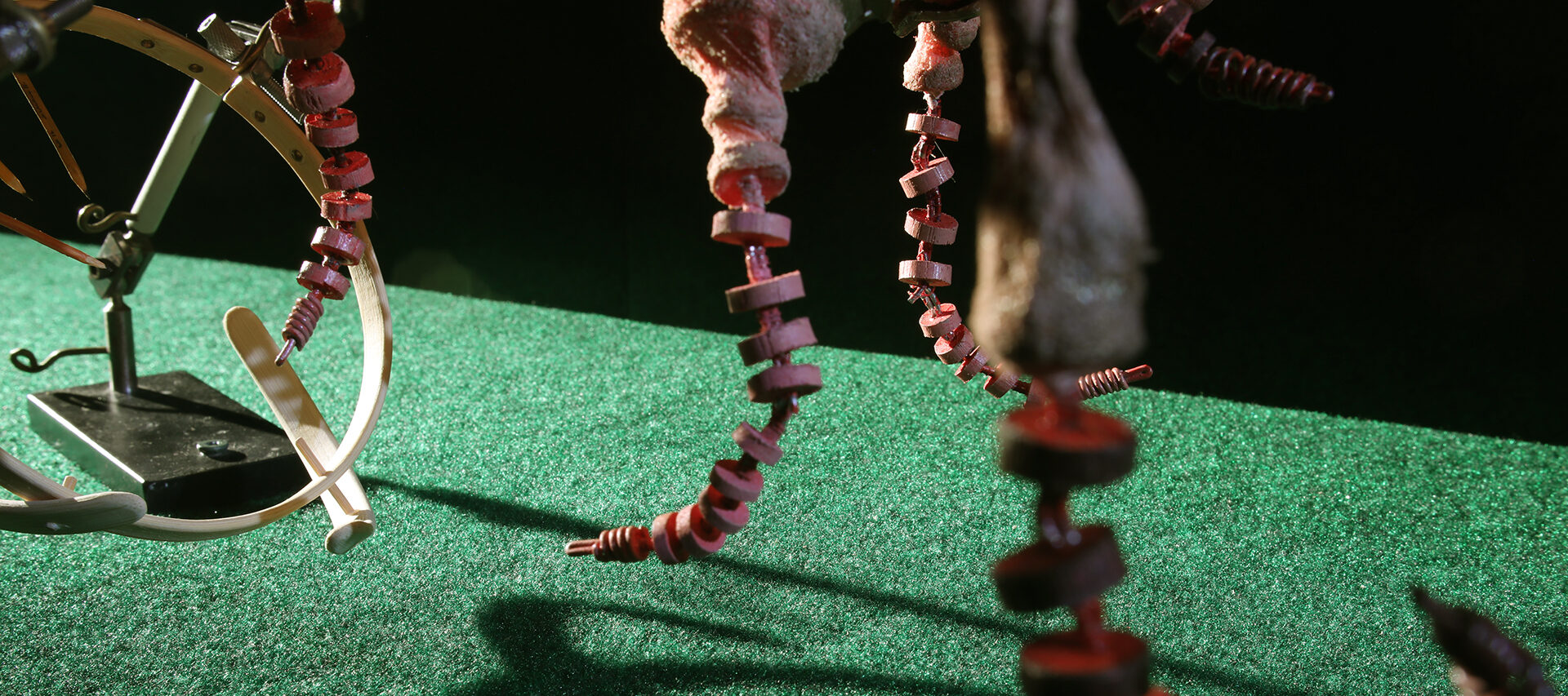 In this screen still from a puppet stop motion animation, minature sculptures made from wood, metal, and other materials are arranged on top of, and hang above, green turf, set against a pitch black background. The sculpture's shadows appear on the turf. The sculptures are made up of various circular designs.