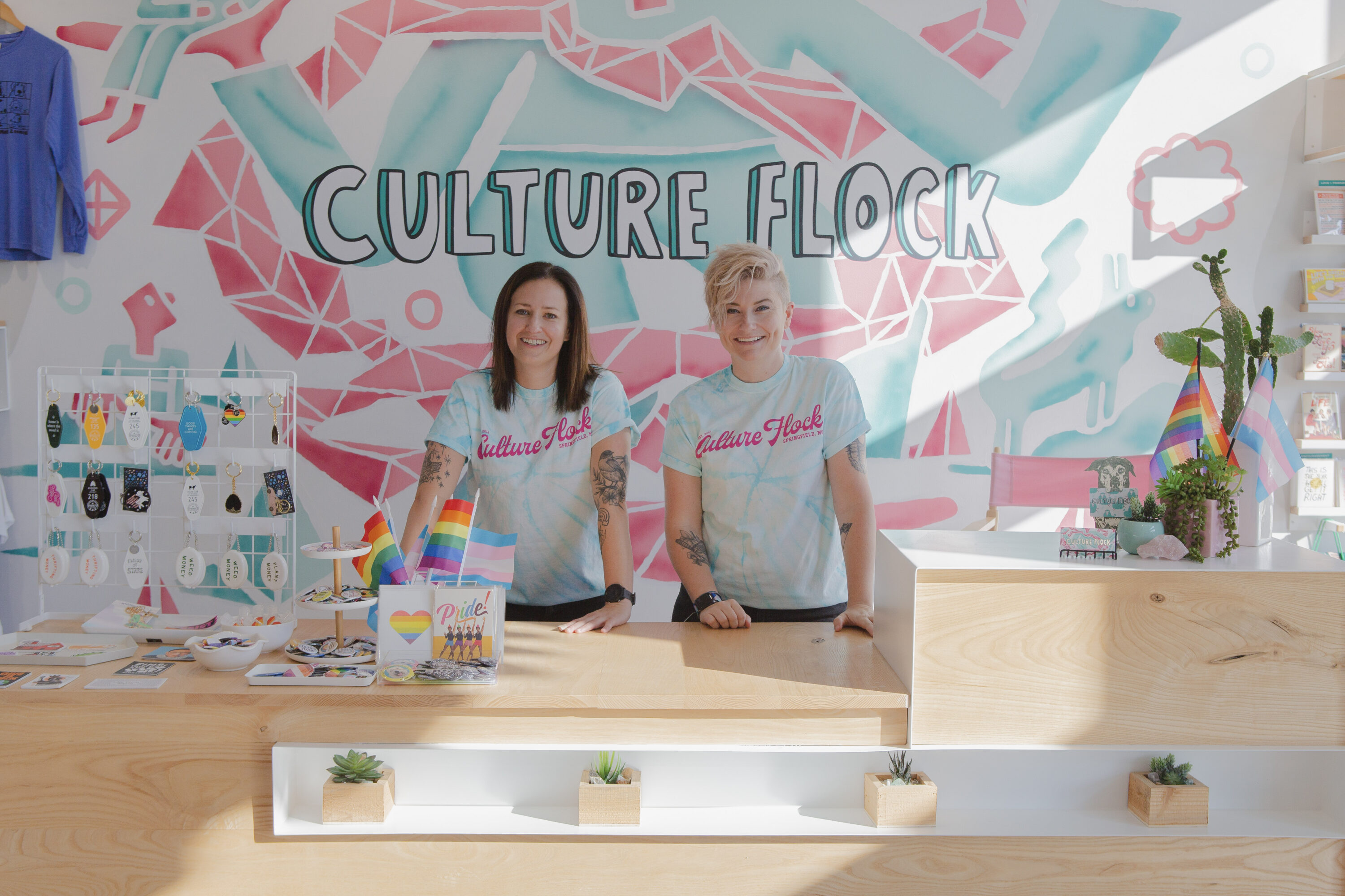 Two light-skinned women stand behind the light-wood counter of a modern retail space. They wear light-blue tie-dyed T-shirts that say "Culture Flock" in bright pink cursive lettering. Both women have tattoos on each arm. They smile brightly. Behind them, "Culture Flock" is written in big, illustrated bubble letters on the wall against a light blue and pink geometric background. On the counter, a display of colorful keychains, pride flags, and colorful cards and pins are arranged.