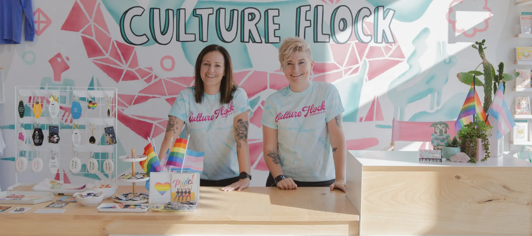 Two light-skinned women stand behind the light-wood counter of a modern retail space. They wear light-blue tie-dyed T-shirts that say "Culture Flock" in bright pink cursive lettering. Both women have tattoos on each arm. They smile brightly. Behind them, "Culture Flock" is written in big, illustrated bubble letters on the wall against a light blue and pink geometric background. On the counter, a display of colorful keychains, pride flags, and colorful cards and pins are arranged.