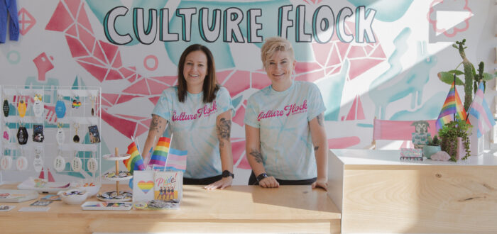 Two light-skinned women stand behind the light-wood counter of a modern retail space. They wear light-blue tie-dyed T-shirts that say "Culture Flock" in bright pink cursive lettering. Both women have tattoos on each arm. They smile brightly. Behind them, "Culture Flock" is written in big, illustrated bubble letters on the wall against a light blue and pink geometric background. On the counter, a display of colorful keychains, pride flags, and colorful cards and pins are arranged.