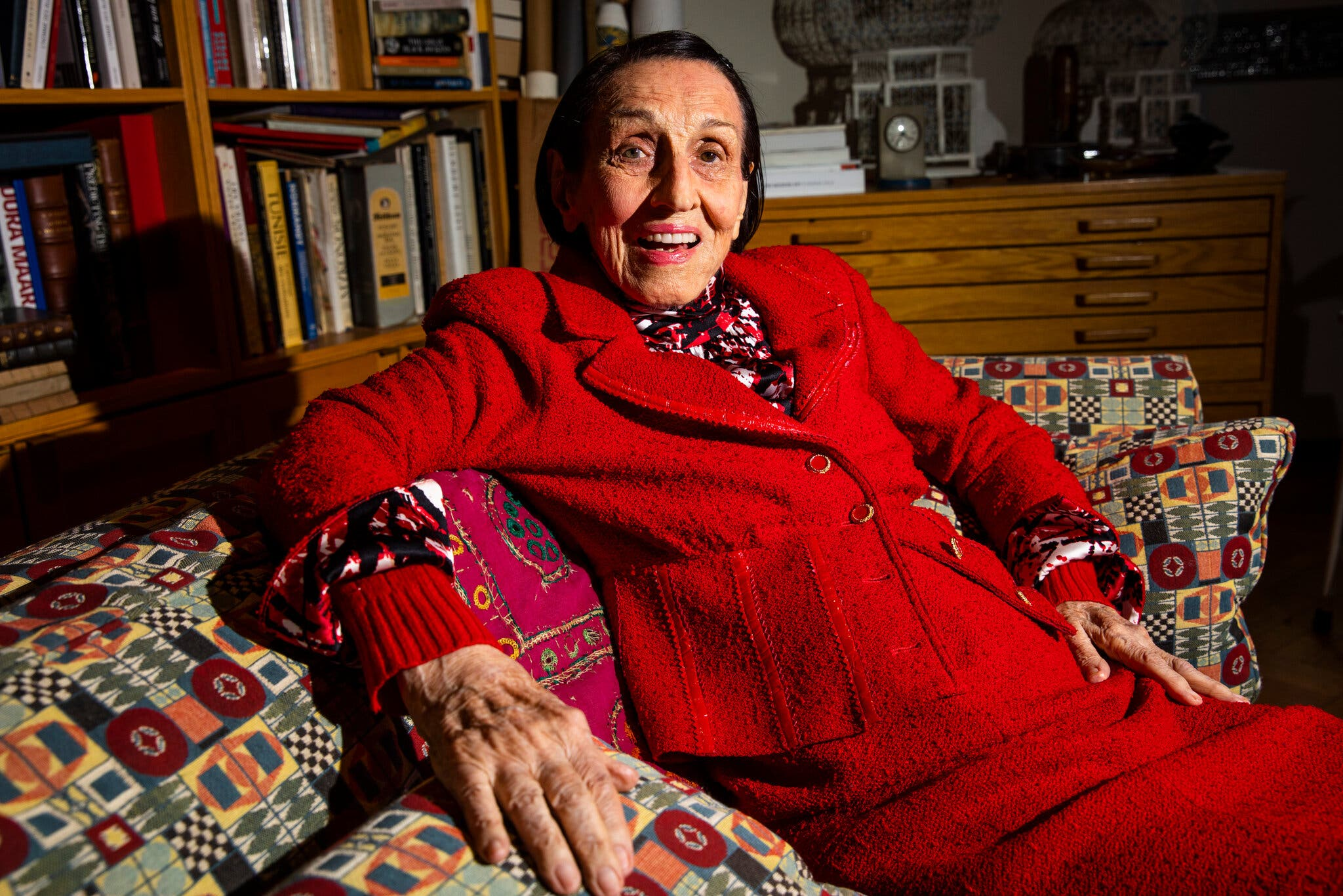 An older, light-skinned woman sits comfortably on a couch covered in geometric print. She wears a bright red skirt and jacket set, and a silk shirt under it with a red, white, and black abstract pattern. Her hair is black, straight, and falls to her earlobes. It is clipped back neatly. She stares at the camera with an enthusiastic look on her face, maybe mid-sentence or mid-smile. Behind her are full bookshelves and a dresser with a clock, and more books.