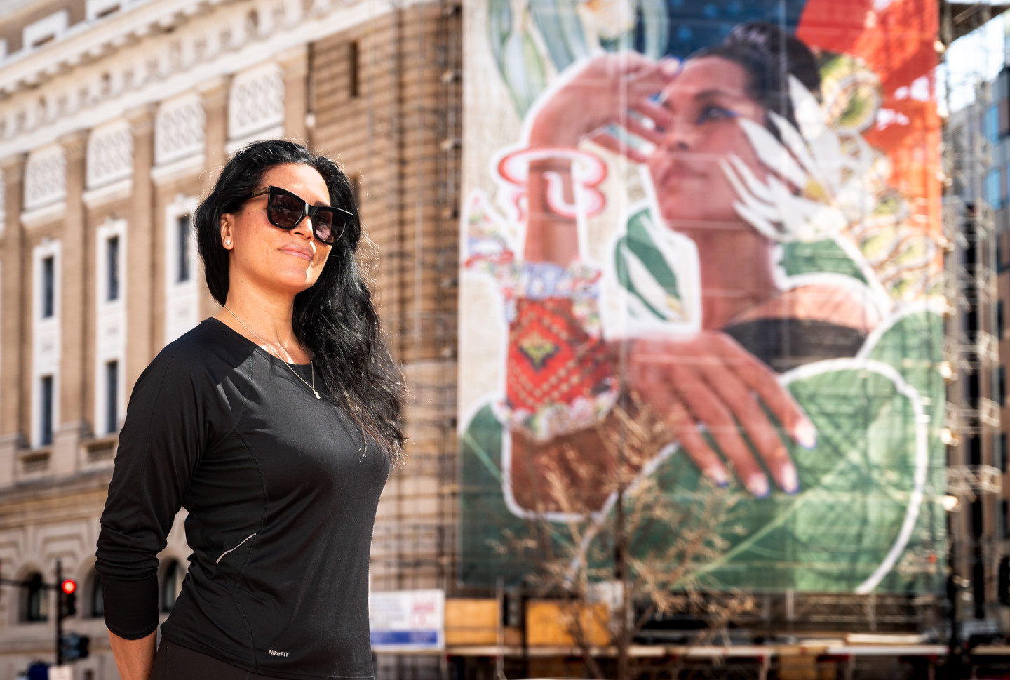 A woman with medium skin tone with long dark hard, sunglasses, and a black shirt stands in front of the exterior of the museum and the large, colorful mural on the front.