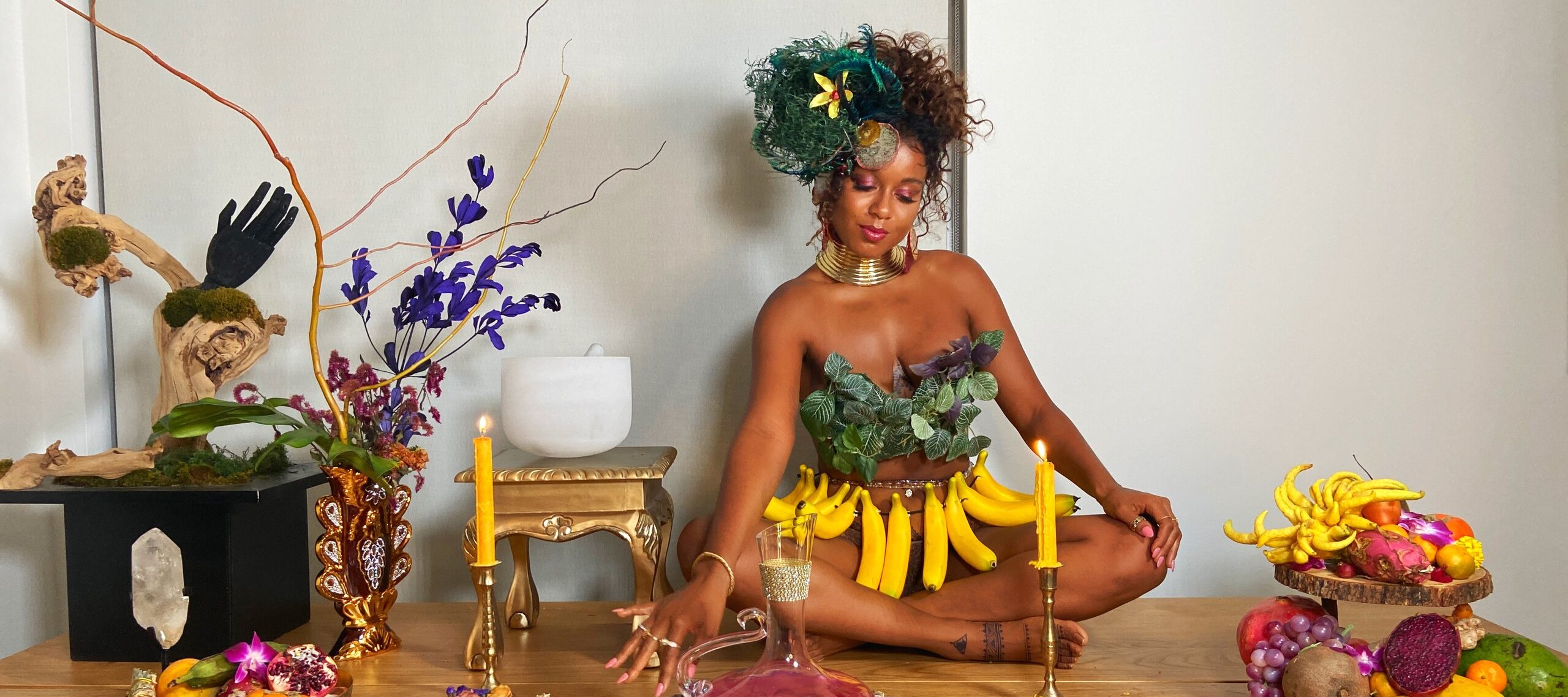 A medium-dark skinned adult woman sits cross-legged on a long wooden table set with platters of colorful fruit, candles, plants, and a pitcher of a pink liquid. She wears green leaves on her chest, gold jewelry, a skirt of bananas, and her hair pulled up with green feathers.