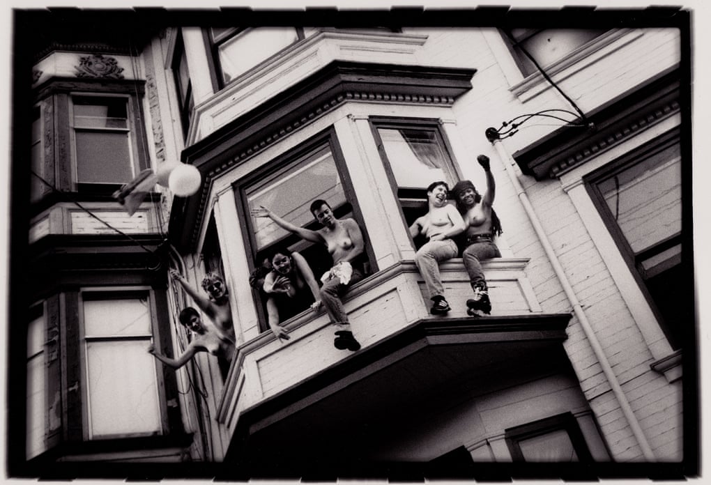 In this black-and-white photo, five topless women emerge from three bay windows in a San Francisco row house. Most have short, cropped hair and are light-skinned. There is one dark-skinned woman with dreadlocks. They smile, laugh, and raise their arms/fists as they look down, seemingly at something happening on the street below.