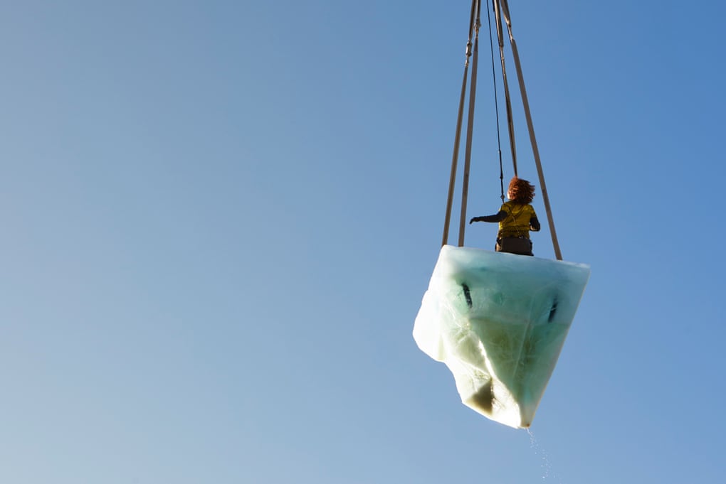 Against a clear blue sky, a white iceberg in the shape of a diamond is suspended by four dark cords. In the center of it, a woman stands with her back to the camera, also suspended by a dark cord. Her left arm is raised slightly.