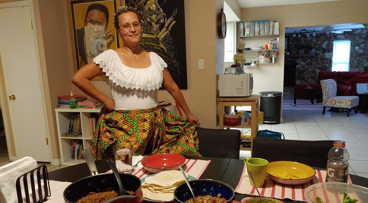 A medium skin-toned adult woman wearing traditional Puerto Rican dress—a white blouse with lace neck and brightly patterned skirt—stands smiling in front of her dining table in her home. The table is set with bowls and pots of ground beef, salsa, sauce, guacamole, and salad.