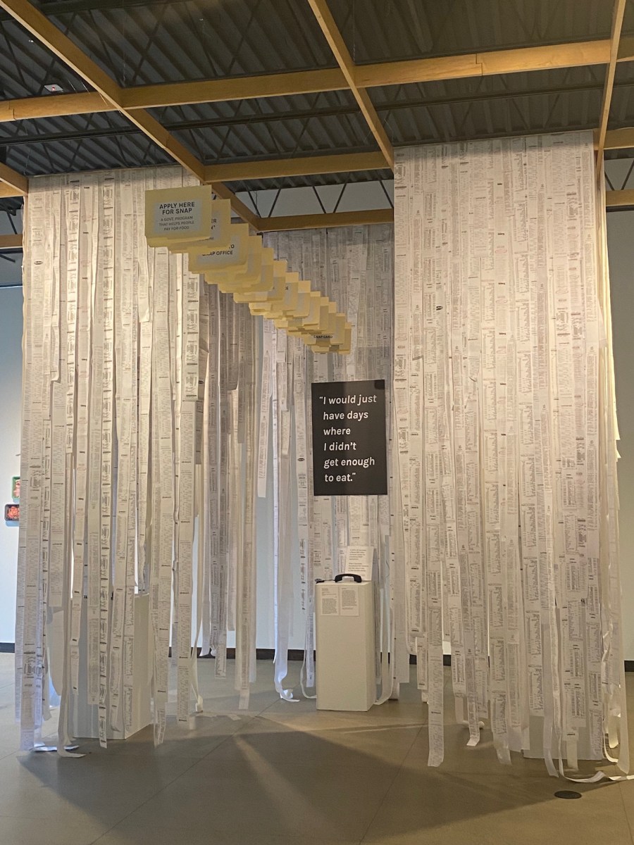 In an industrial gallery space, reams and reams of, seemingly, store reciepts hang like curtains from the tall ceiling, some grazing the ground. In the middle of this is a suspended sign that says "I would just have days where I didn't get enought to eat." Below it is a podium with a phone and various pages of text, unreadable at the distance the photo is taken.