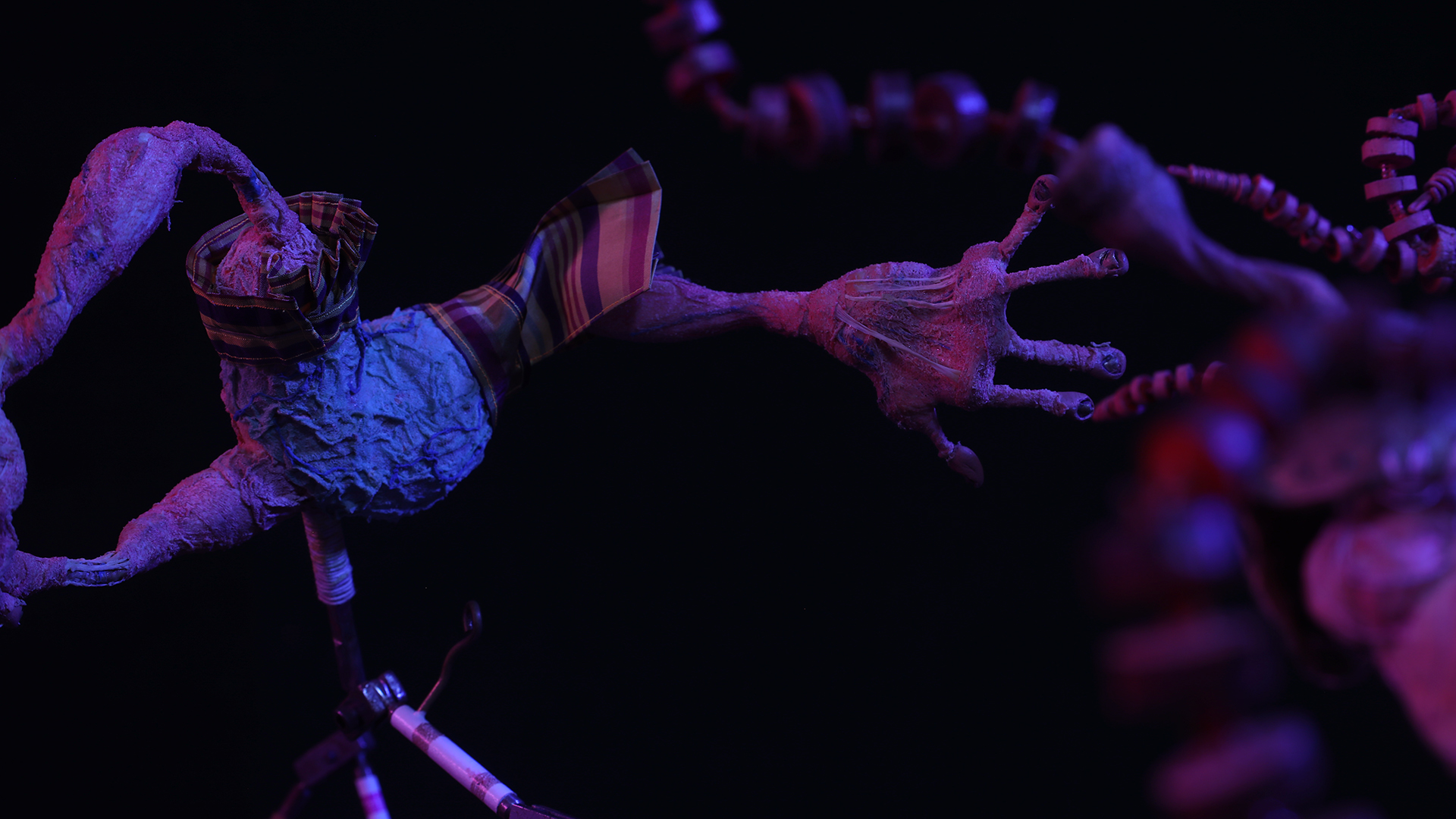 In this screen still from a puppet stop motion animation, a creepy abstract form contains a hand reaching out toward the right. It is made from a dehydrated-looking, wrinkled material and the hand is misshapen, but does contain five fingers. A patch of plaid fabric sits on the shoulder and is tied around the "neck," which just contains a long, strange tube emerging from it and made from the same material.