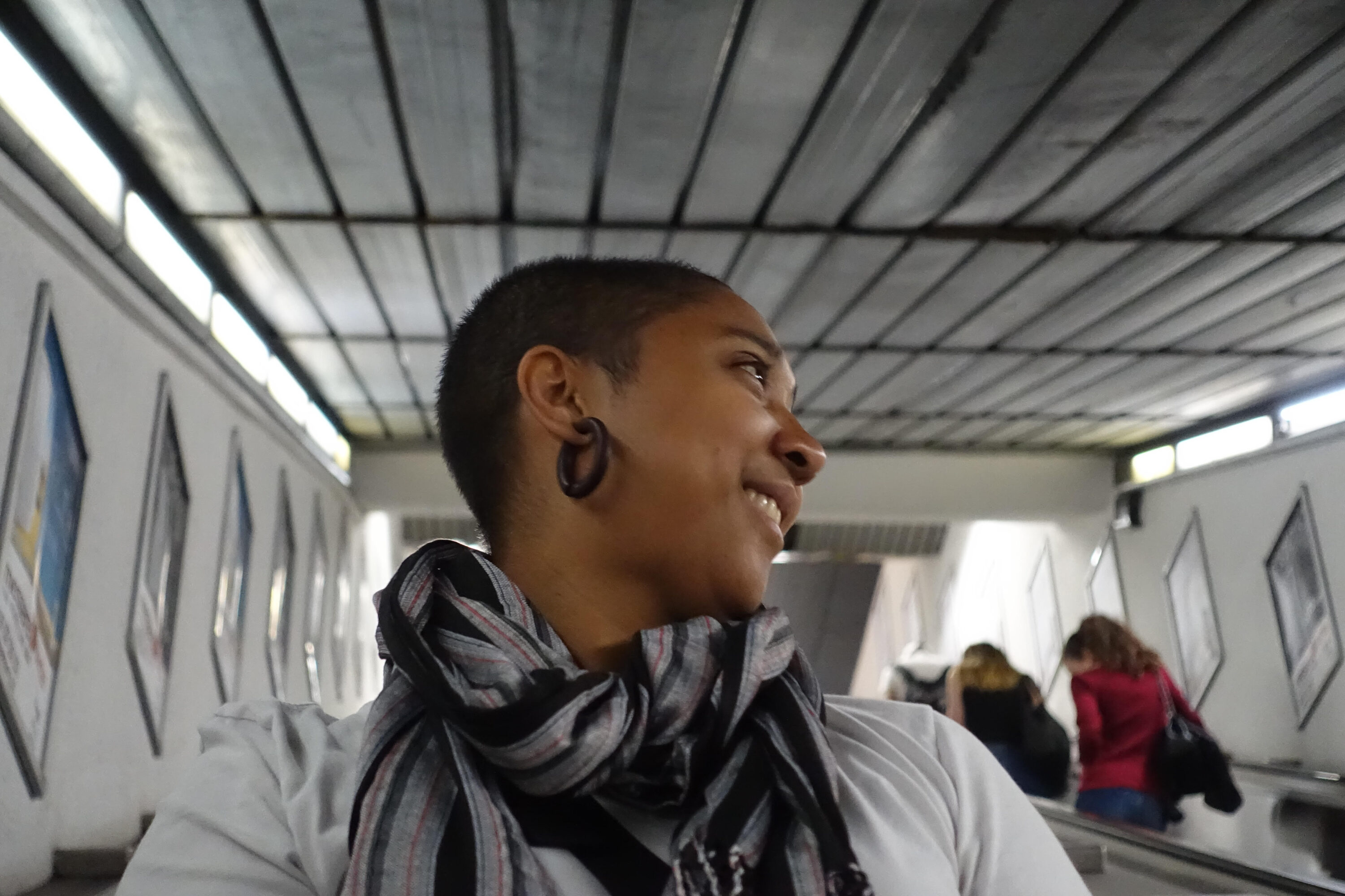 A portrait photo of a dark-skinned woman with a shaved head looking off to the right and smiling. She wears a black, white, and red striped scarf tied around her neck atop a white shirt.