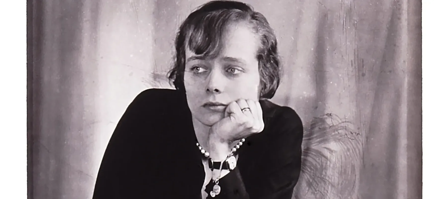 A black-and-white photograph of a light-skinned woman with short cropped hair leaning on a table with her left hand holding her chin. She looks off to the left, unsmiling.