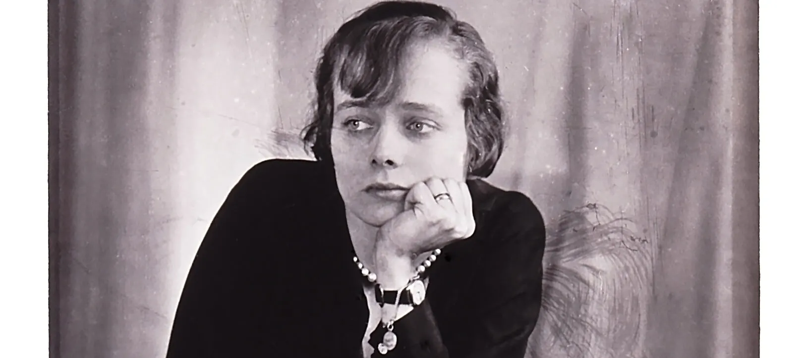 A black-and-white photograph of a light-skinned woman with short cropped hair leaning on a table with her left hand holding her chin. She looks off to the left, unsmiling.