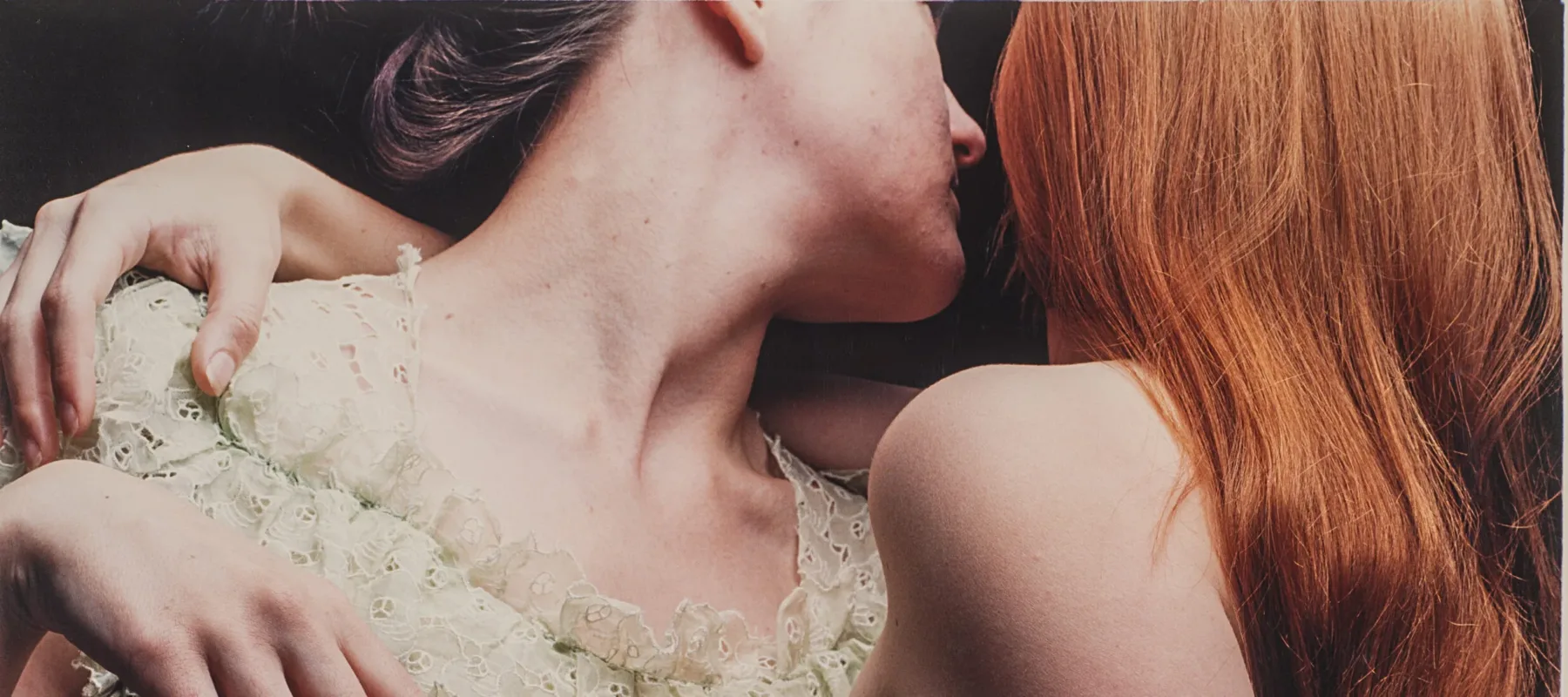 Two light-skinned people, one wearing a lace dress and the other nude, with their arms around each other and facing away from the viewer. The person on the left has brown hair pulled back and the person on the right has long, red hair.