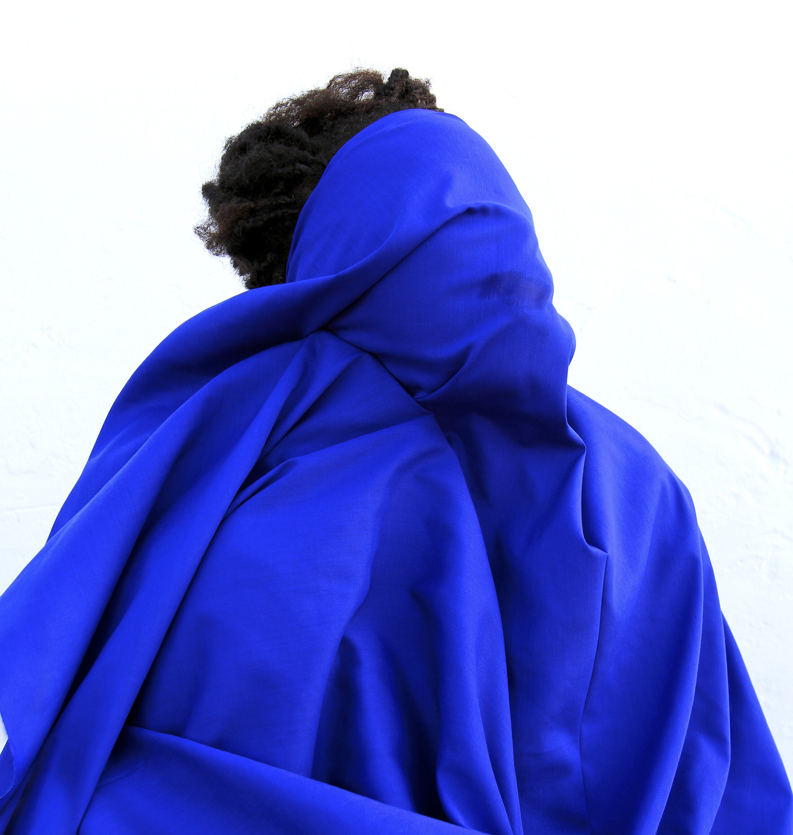 A figure with short dark hair stands in front of a stark white background with voluminous, vibrant blue fabric draped over their face and body.