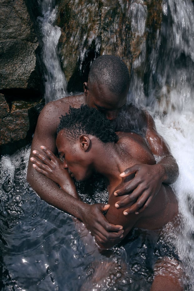 A photograph of two naked dark-skinned men at the base of a small waterfall. The embrace tenderly as the water rushes behind them and into the glistening pool that surrounds them.