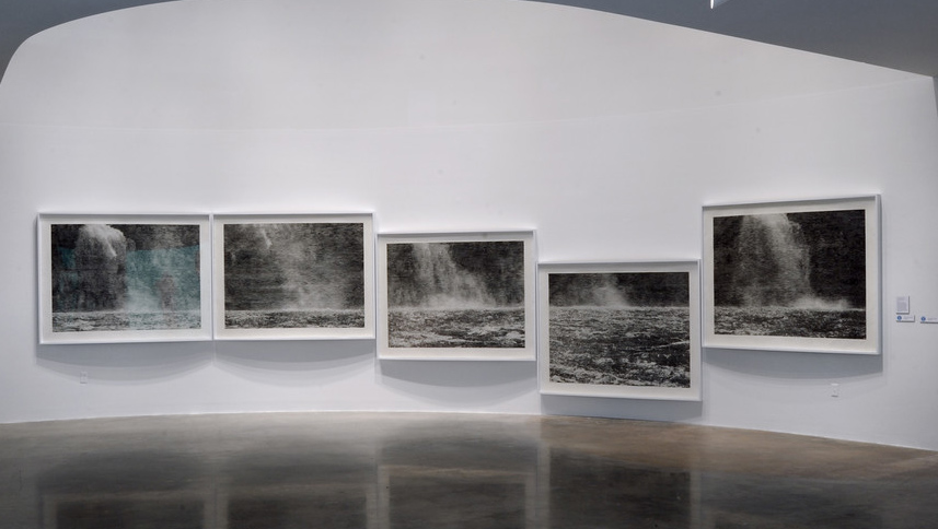 A series of five black and white prints hang on a white wall. The prints depict different sections of a panoramic view of a waterfall and the pool of water below it. Two of the five prints are hung lower than the rest but the line where the pool meets the waterfall remains consistent across all five prints.