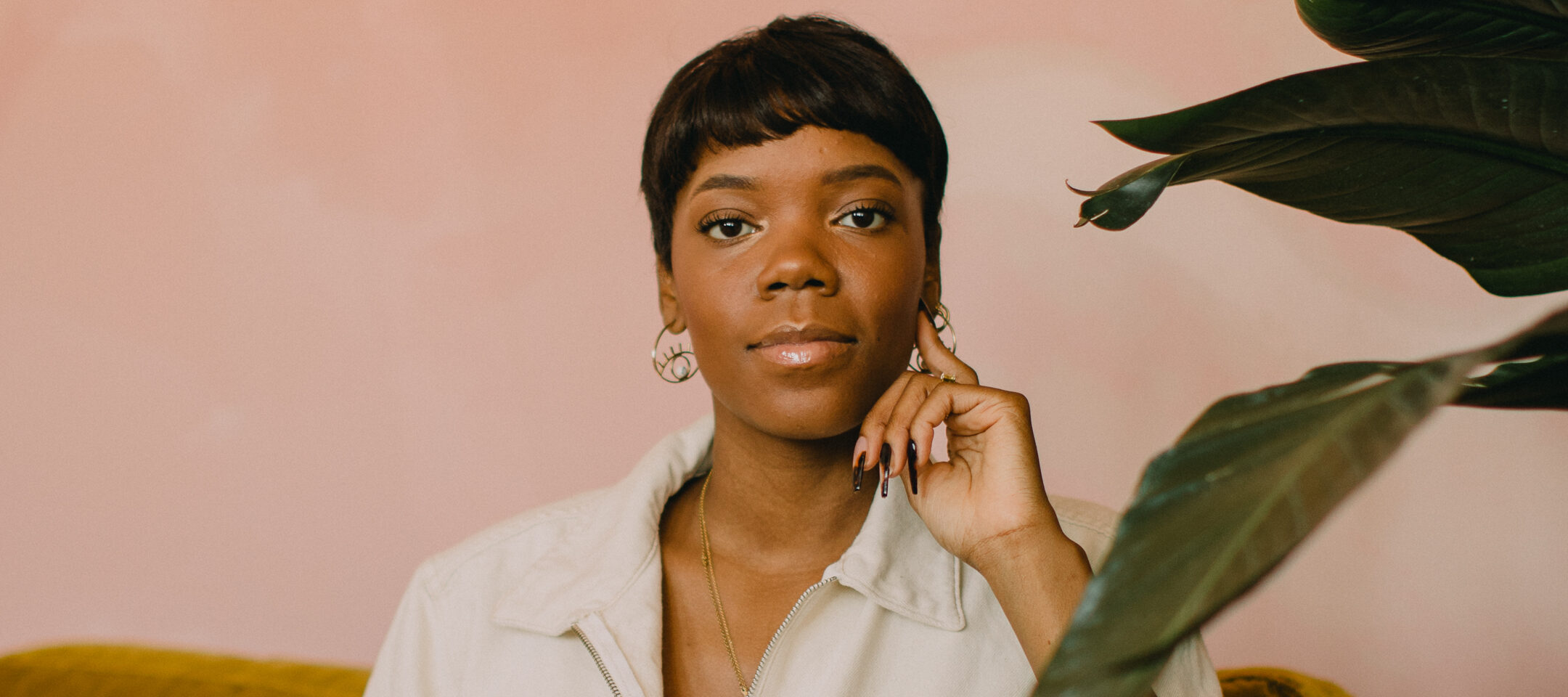 A dark-skinned woman with a short, straight pixie cut sits in a relaxed pose on a velvet mustard-colored couch. She wears a off-white jumpsuit and raises her left hand to rest on her cheek as she smiles slightly at the camera. The wall behind her is painted a blush-pink color and a wide-leaf large plant is situated to her right.