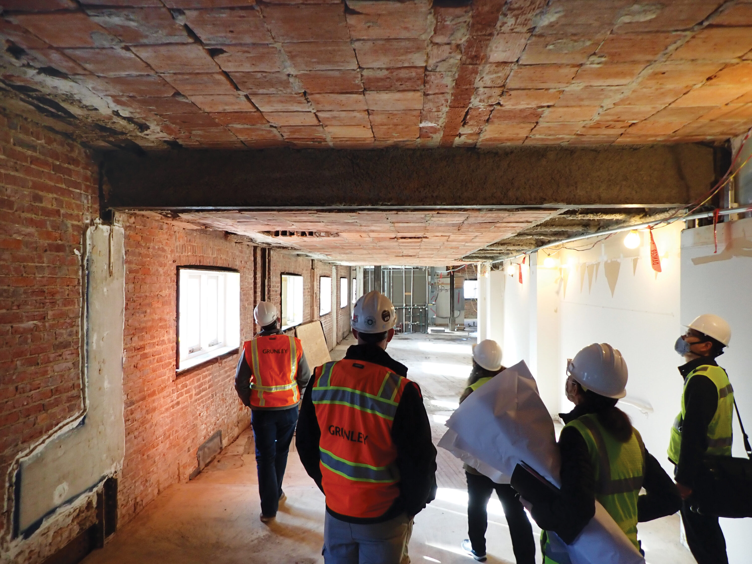 A team of five construction workers stand in the hallway of the museum building as it is under renovation. They wear white hard hats and yellow and orange safety vests over business casual clothes. They look up and down the hall, which is stripped down to it's base layer of red brick, concrete, and thick beams.