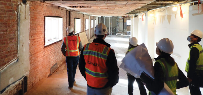 A team of five construction workers stand in the hallway of the museum building as it is under renovation. They wear white hard hats and yellow and orange safety vests over business casual clothes. They look up and down the hall, which is stripped down to it's base layer of red brick, concrete, and thick beams.