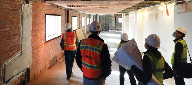 A team of five construction workers stand in the hallway of the museum building as it is under renovation. They wear white hard hats and yellow and orange safety vests over business casual clothes. They look up and down the hall, which is stripped down to it's base layer of red brick, concrete, and thick beams.