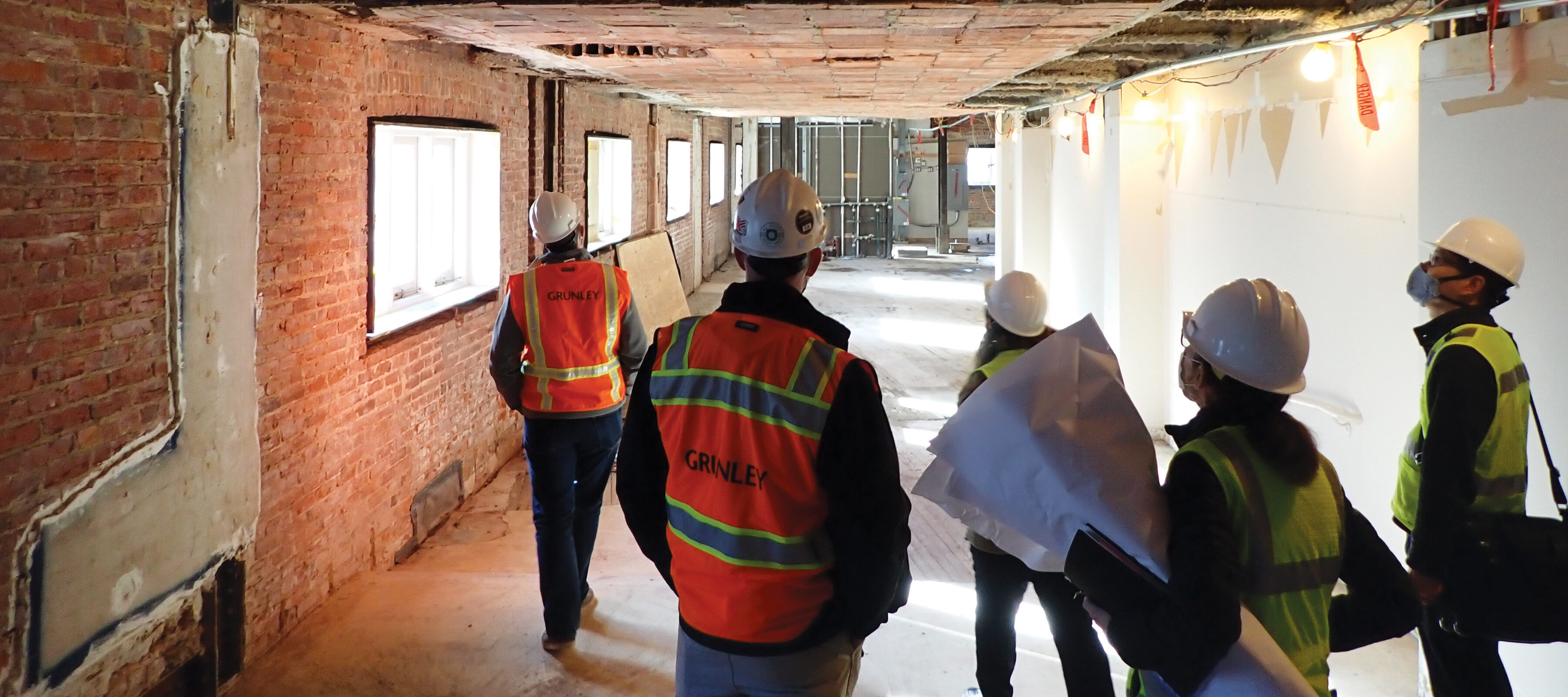 A team of five construction workers stand in the hallway of the museum building as it is under renovation. They wear white hard hats and yellow and orange safety vests over business casual clothes. They look up and down the hall, which is stripped down to it's base layer of red brick, concrete, and thick beams.