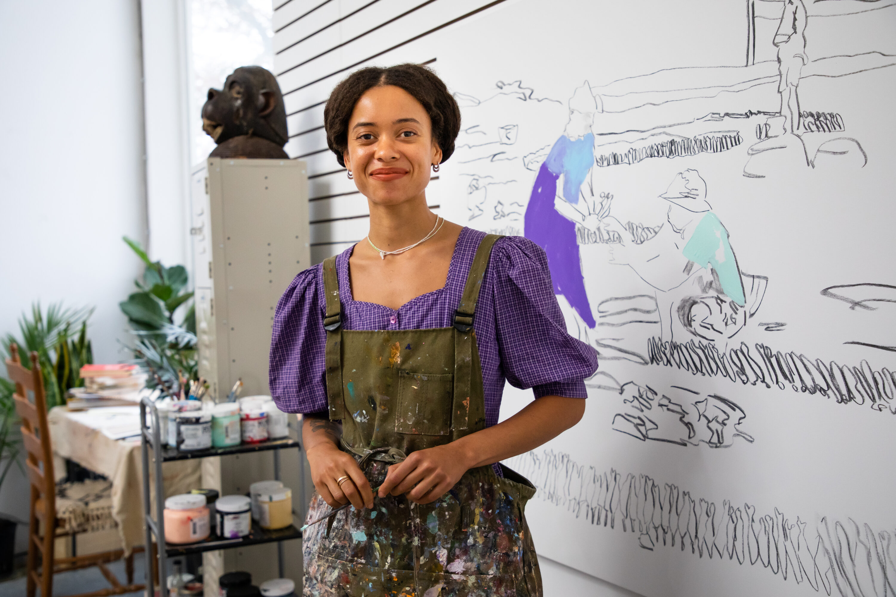 A medium-skinned woman with brown hair that is parted in the middle and pulled back, stands in an artist studio wearing a short-sleeved purple blouse and olive green overalls covered in paint. Behind her is an unfinished painting of three figures outlined in black with highlights of purple, blue and green.