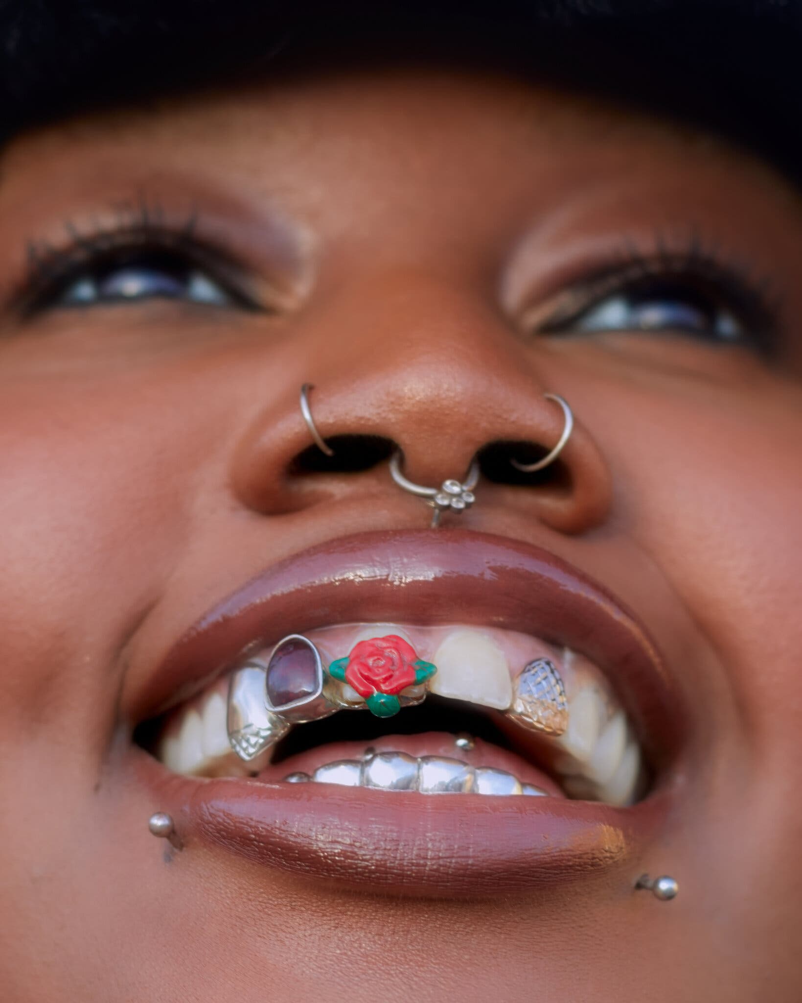 A close-up portrait of a dark-skinned woman, focused on her mouth. She wears multiple, intricate grills over her teeth. One is a red rose with green leaves, the others are different light colors or silver. She smiles widely to show off the pieces and has three nose and two lower-lip piercings. Her lips are perfectly glossed in a coffee color.