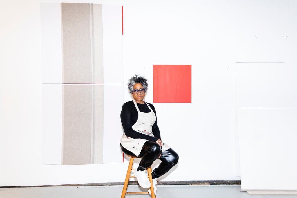 A dark-skinned woman with a salt-and-pepper afro sits on a low stool in a modern studio space. She wears shiny black pleather pants, a black long sleeved shirt and a white apron over both. Behind her hands a large painting featuring a coral square in its center, surrounded by white.