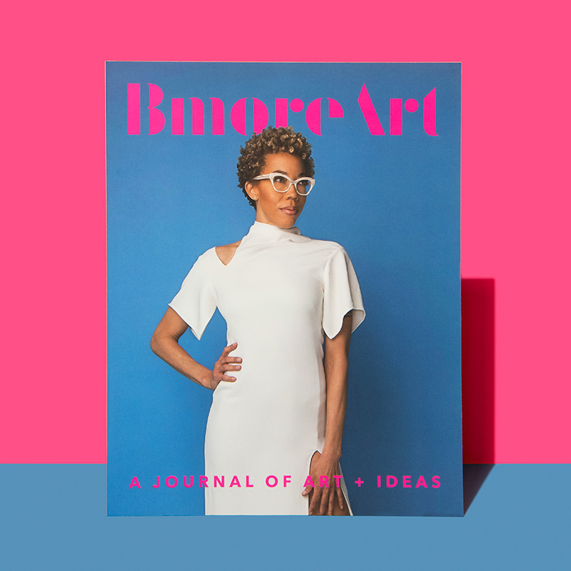 A magazine is posed in front of a bubblegum-pink wall and baby blue base. The magazine itself features a dark-skinned woman with a short afro standing in front of a blue background in an elegant white cocktail dress. Above her the words "BmoreArt" are arranged prominently in pink. Below reads "A journal of art + ideas."