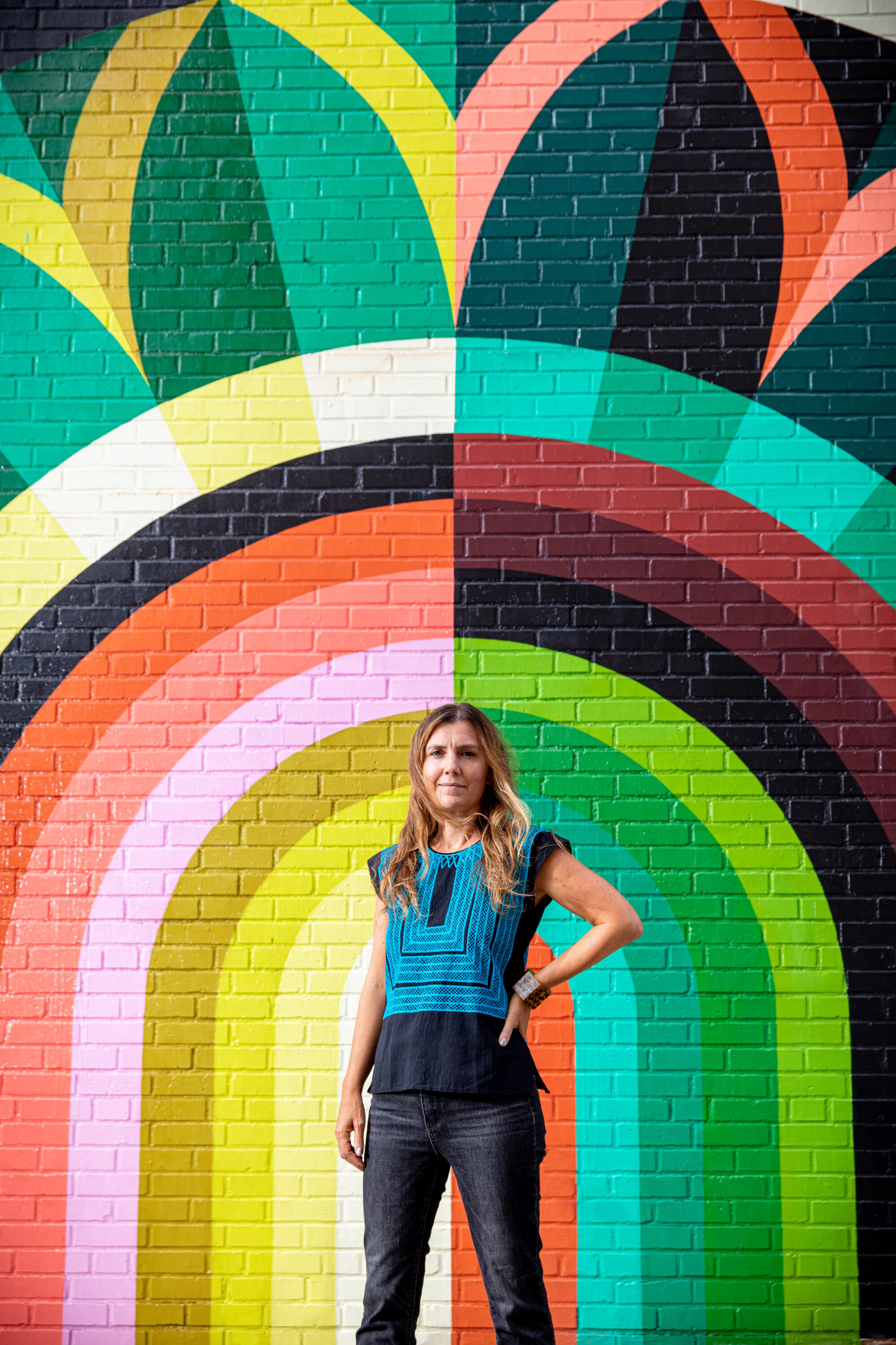 A light-skinned woman with dirty blonde, straight hair stands in front of a large colorful mural painted atop a brick wall. It features rainbow-like stripes in different muted hues, atop which petal shapes are arranged like a crown. The woman stands straight and erect, her left hand on her hip.