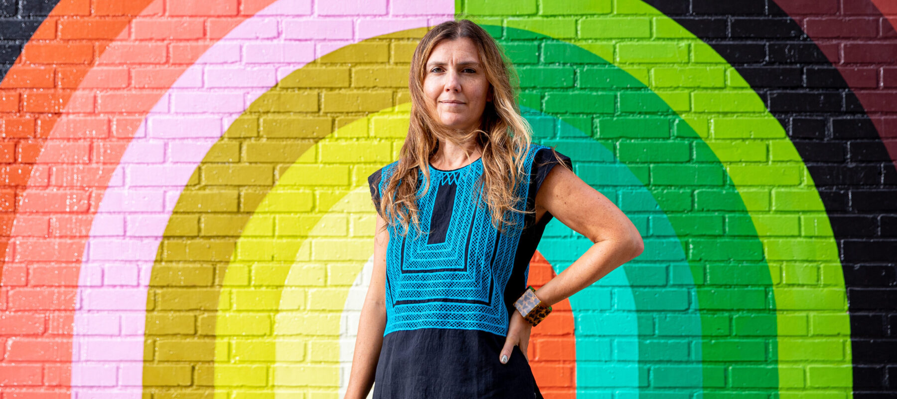 A light-skinned woman with dirty blonde, straight hair stands in front of a large colorful mural painted atop a brick wall. It features rainbow-like stripes in different muted hues, atop which petal shapes are arranged like a crown. The woman stands straight and erect, her left hand on her hip.