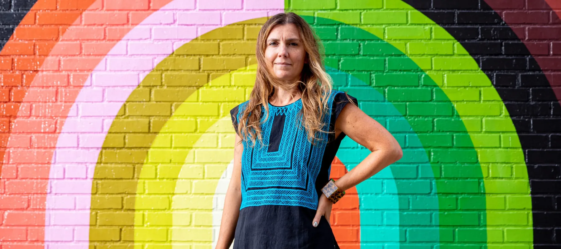 A light-skinned woman with dirty blonde, straight hair stands in front of a large colorful mural painted atop a brick wall. It features rainbow-like stripes in different muted hues, atop which petal shapes are arranged like a crown. The woman stands straight and erect, her left hand on her hip.