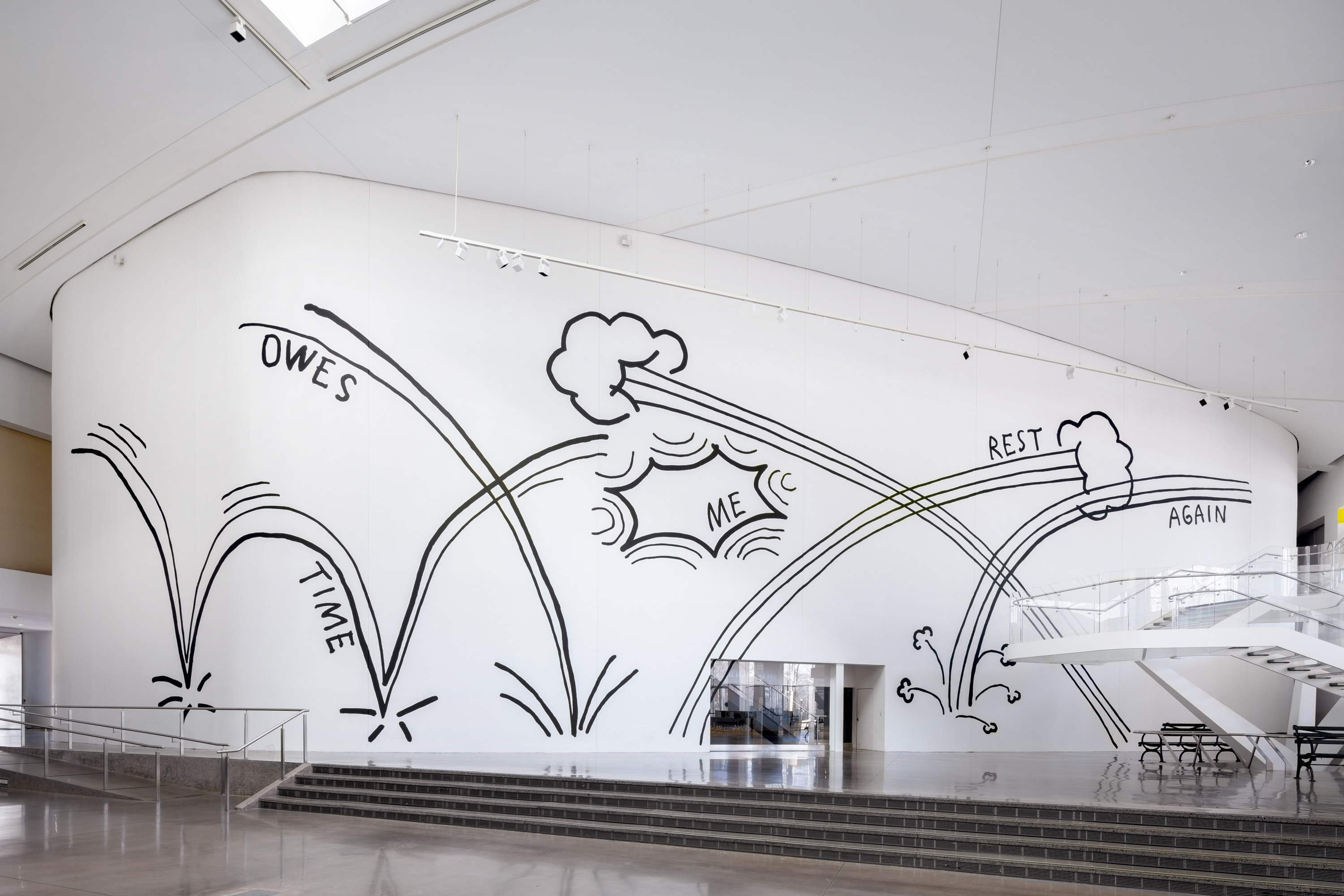 A photo is taken from a distance to show a large mural painted on a white wall. The mural is done in black paint and features the phrase "Time Owes Me Rest Again" arranged on comic book-esque action lines, which bounce and zip across the wall.