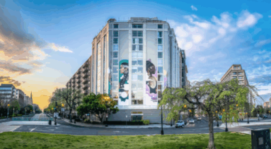 On the side of a building, a six-story mural features two women in profile, holding spears and looking out. The woman on the left has medium-light skin tone, a long dark and gray braid, and silver earrings while the woman on the right has dark skin tone, a dark bun, and a gold breastplate