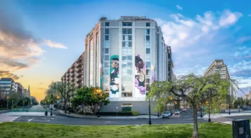 On the side of a building, a six-story mural features two women in profile, holding spears and looking out. The woman on the left has medium-light skin tone, a long dark and gray braid, and silver earrings while the woman on the right has dark skin tone, a dark bun, and a gold breastplate