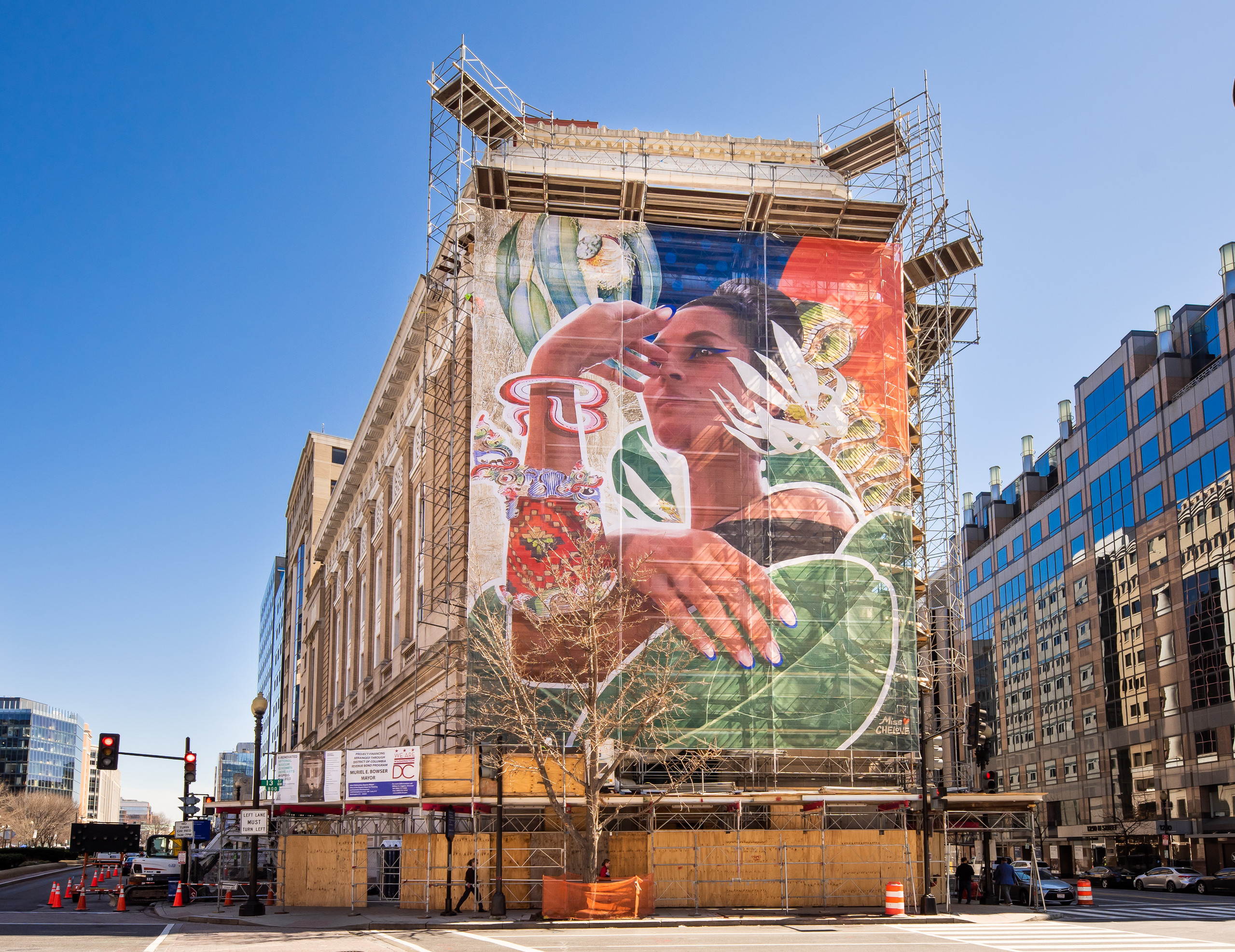 One side of a city building is covered in scaffolding as it is under construction. The scaffolding is covered with a scrim, featuring a digital collage artwork of a woman’s head and shoulders that are partially obscured by leaves, flora, and abstract graphics. She has medium-light skin tone and black hair worn in a high bun, and she looks up and to the side in a regal pose.