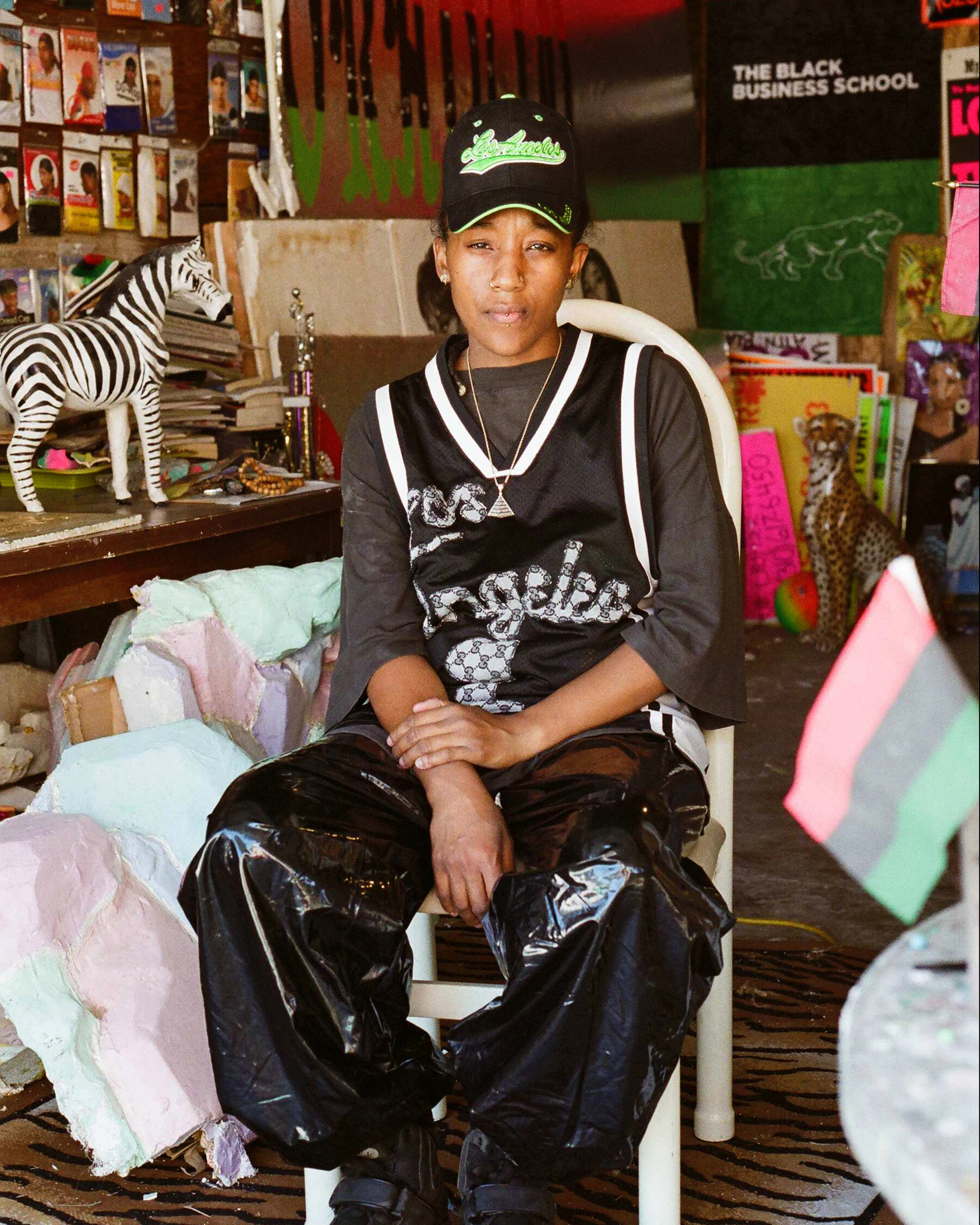 A dark-skinned woman sits in a white chair in what looks to be a busy artist's studio space. It is filled with posters, canvases, materials, a zebra figurine on a desk, and a cheetah sculpture on the floor in the far back. The woman wears a baseball hat, sports jersey over a grey shirt and shiny, baggy, black pants.