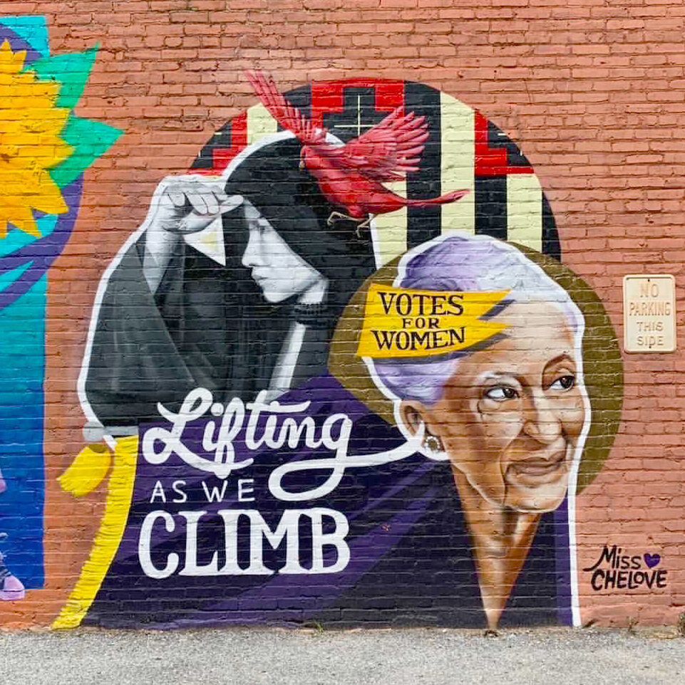 The head of an older woman with medium skin tone and white hair that is partially obscured by a yellow flag that says “votes for women.” Behind her, a black-and-white figure wears a black, hooded cloak by a red bird. “Lifting as we climb” is painted in white against a dark purple background.