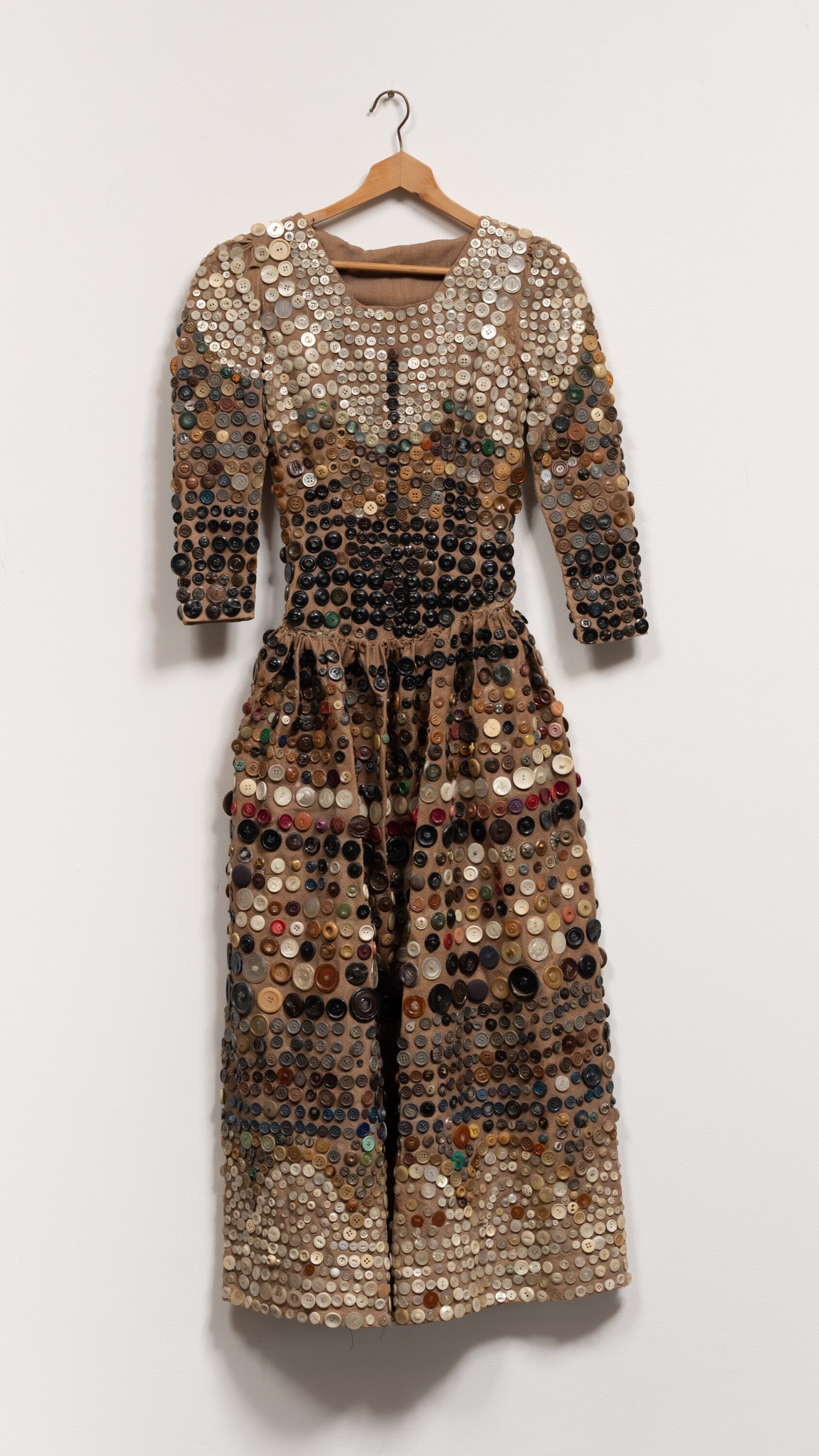 A dress made from earth-toned buttons hangs on a wooden hanger on a white gallery wall. The sleeves are three-quarter length and the skirt is long, possibly hitting at the mid-calves. Clear, white, tan, and dark brown and black buttons are arranged in rows and scalloped patterns.