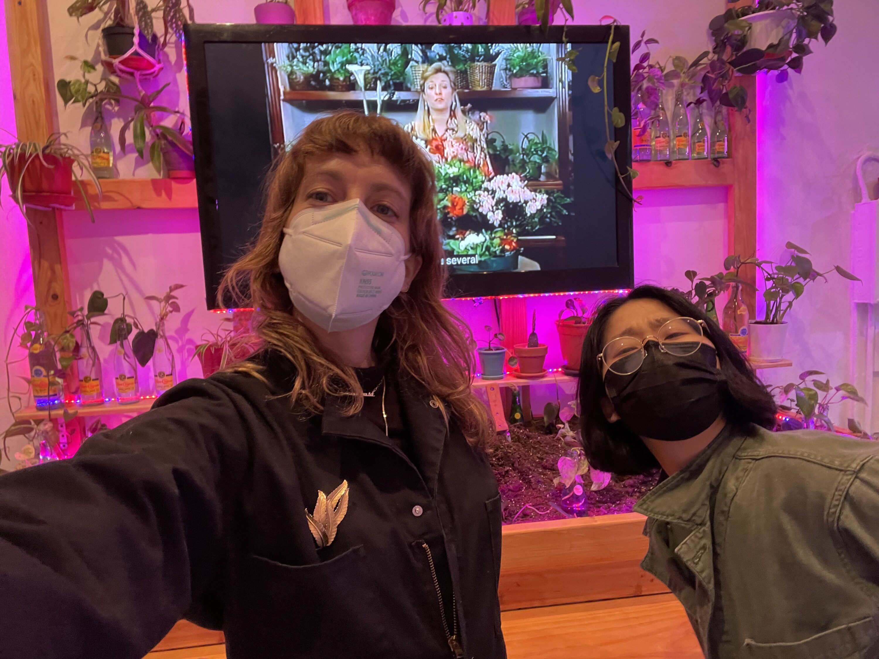 Two women with light skin tones are taking a selfie in front of a wooden consctruct holding plants and pots.